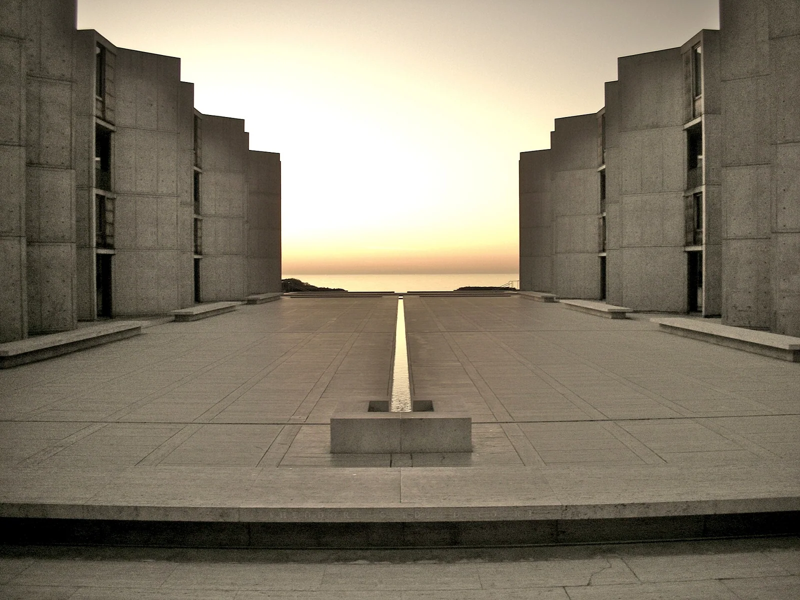 A travertine plaza with a narrow water channel leading toward a sunset horizon between concrete walls.