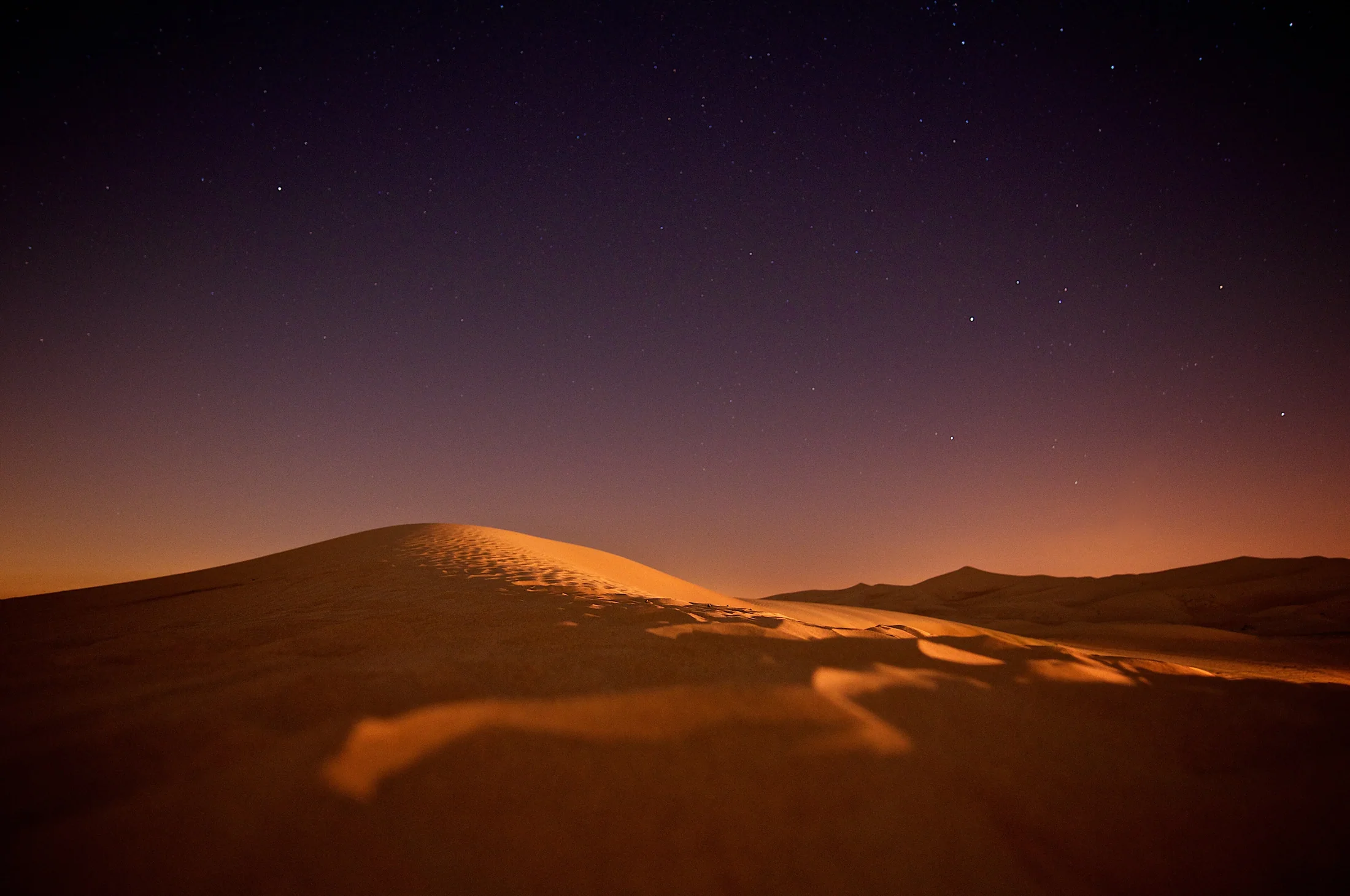 Amber desert dunes under a deepening twilight sky with the first stars appearing.